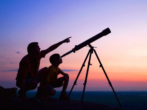 A young boy looking thru a telescope at sunset with his father. - biotimes Vision